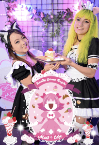 Two women in main uniforms and cat ears are holding a piece of cake in a cute booth with flowers. the sign says "ready Game Begin" and "maid café" with small images of desserts surrounding the sign. 