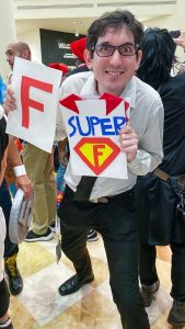 man with glasses is smiling holding a paper in his left hand with a giant F on it. the other hand has another paper with a "super" F and a cape