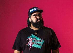 A photo of a person named Awesomus Prime wearing a black graphic T-shirt, glasses, and a black hat in front of a hot pink background